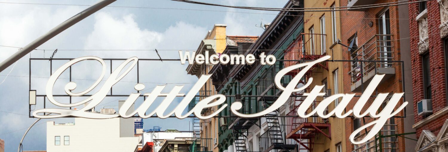 Uma grande placa diz "Bem-vindo à Little Italy" acima de uma rua da cidade com prédios ao fundo. A foto é capa do post Tour gastronômico por Little Italy e Chinatown.