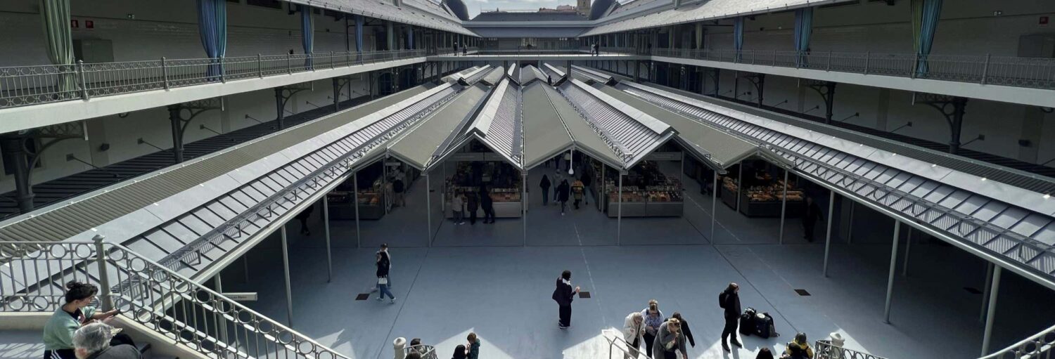 As pessoas caminham e se reúnem em um grande mercado interno com fileiras de barracas de vendedores e um teto alto de vidro. A foto é capa do post Tour gastronômico pelo mercado do Bolhão.