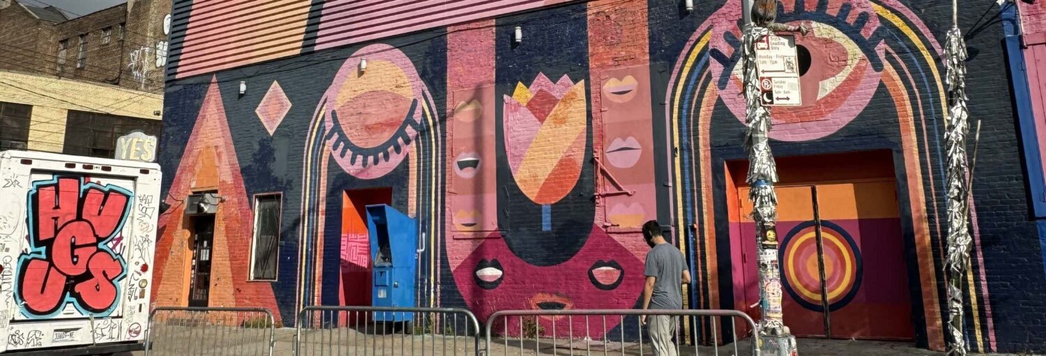Uma pessoa passa por um mural colorido com formas abstratas e rostos em um prédio de tijolos, ao lado de uma van de grafite "HUGS". A foto é capa do post tour do grafite pelo Brooklyn.