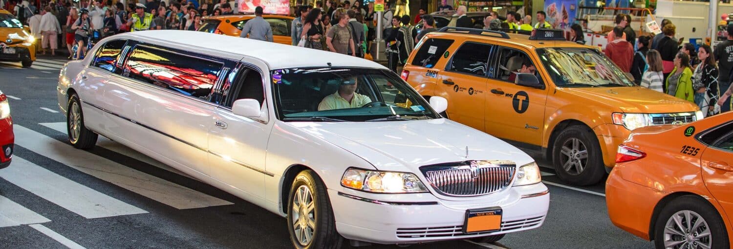 Uma limusine branca passa por uma movimentada rua da cidade, cercada por táxis amarelos e multidões de pedestres. A foto é capa do post de tour privado de limousine por Nova York.