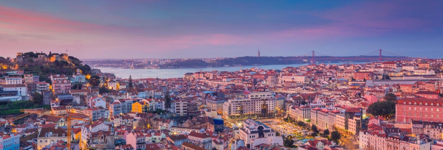 Uma vista panorâmica de Lisboa ao pôr do sol, mostrando edifícios, um castelo no topo de uma colina, o rio e uma ponte suspensa. Imagem para ilustrar post sobre tour por Lisboa completo com ingressos.