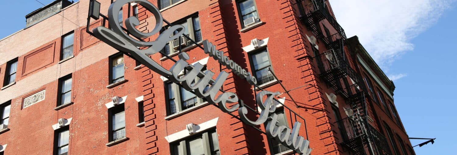 Uma placa de metal "Welcome to Little Italy" está pendurada na frente de um prédio de tijolos vermelhos com escadas de incêndio e janelas. A foto é capa do post de tour por Chinatown e Little Italy.