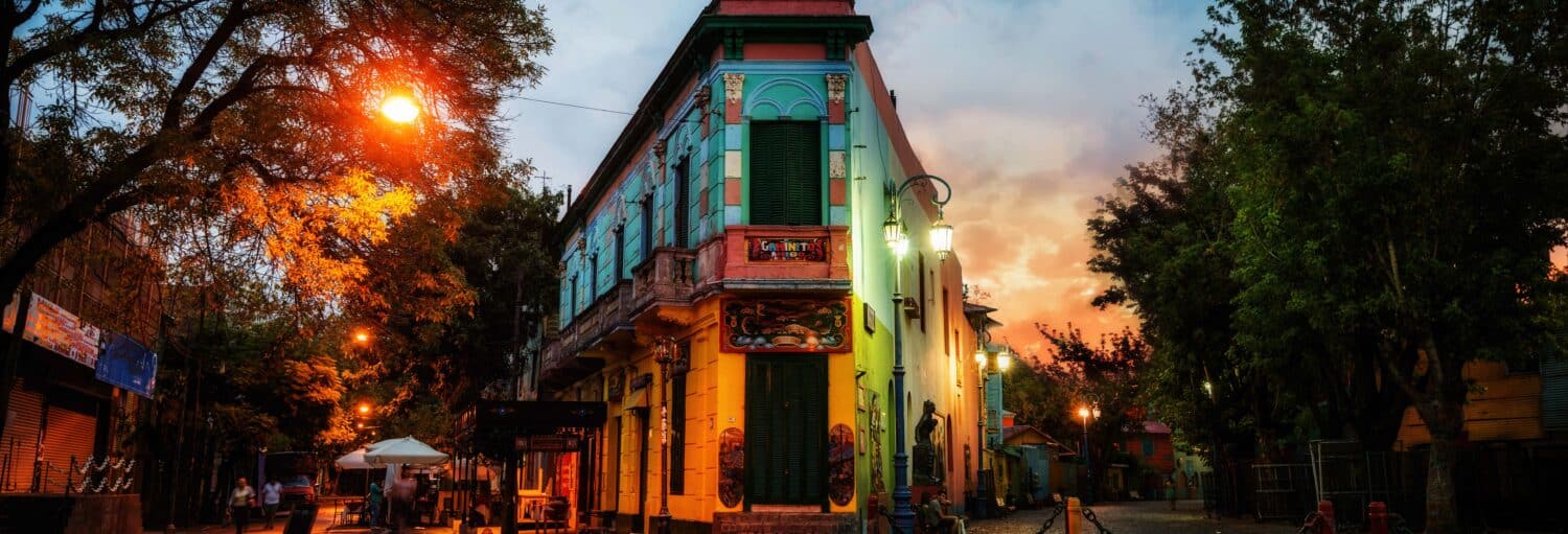 Um edifício colorido fica em uma esquina com árvores e lâmpadas ao pôr do sol. Imagem para ilustrar post sobre tour de mistérios e lendas pelo Sul de Buenos Aires.