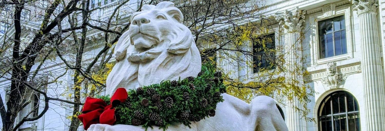 Estátua de leão branco com uma coroa de pinheiros verdes e um laço vermelho em frente a um edifício clássico com colunas e árvores.