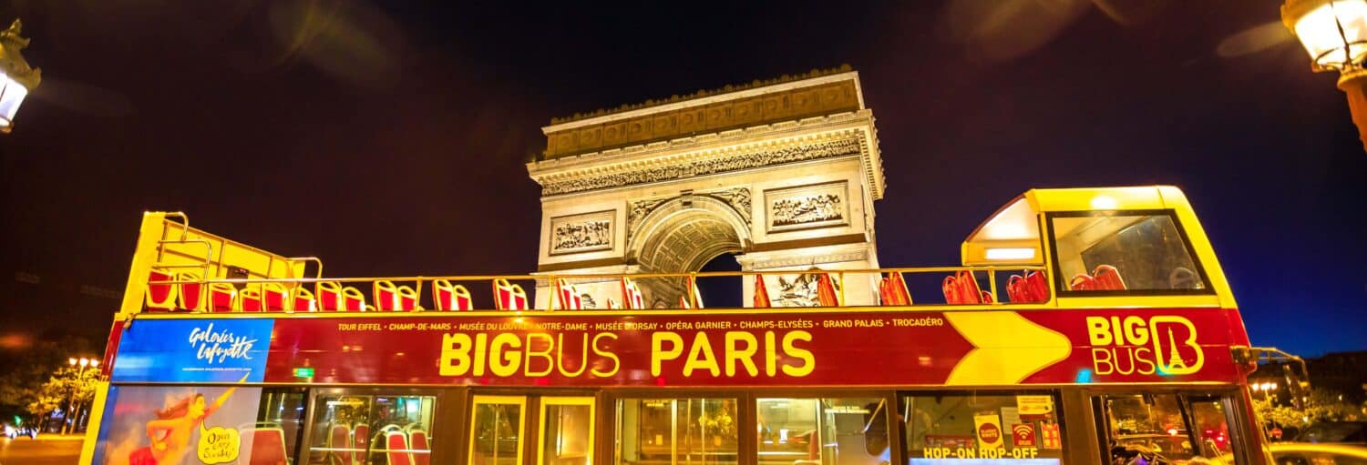 Um ônibus turístico amarelo e vermelho da Big Bus Paris está estacionado em frente ao Arco do Triunfo iluminado à noite. A imagem ilustra o post sobre tour noturno de ônibus panorâmico por Paris.