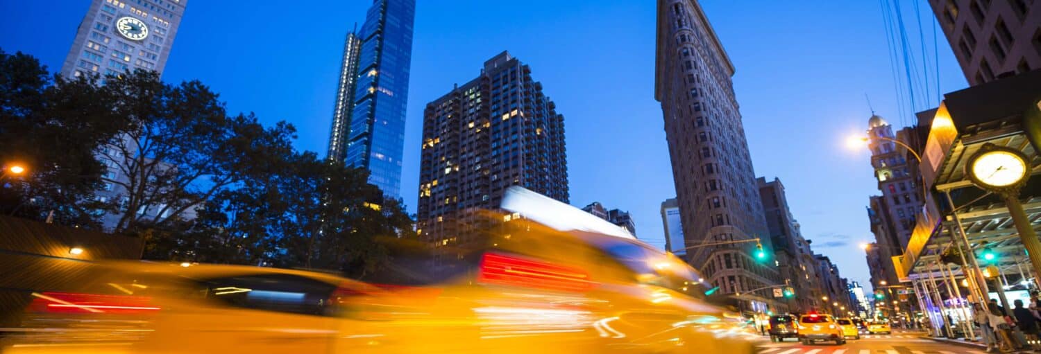 Um táxi amarelo desfocado atravessa uma rua da cidade ao anoitecer, com prédios altos e postes de luz ao fundo. A foto é capa do post tour noturno em ônibus panorâmico.