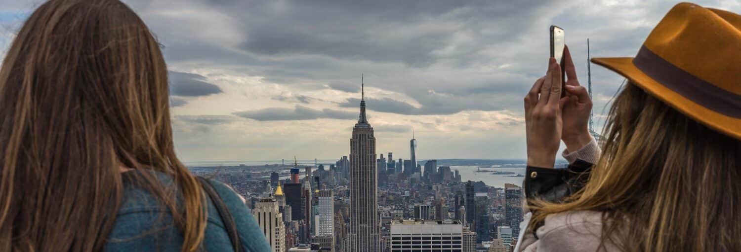 Duas pessoas olham para o horizonte da cidade de Nova York com o Empire State Building; uma pessoa está tirando uma foto com um telefone.