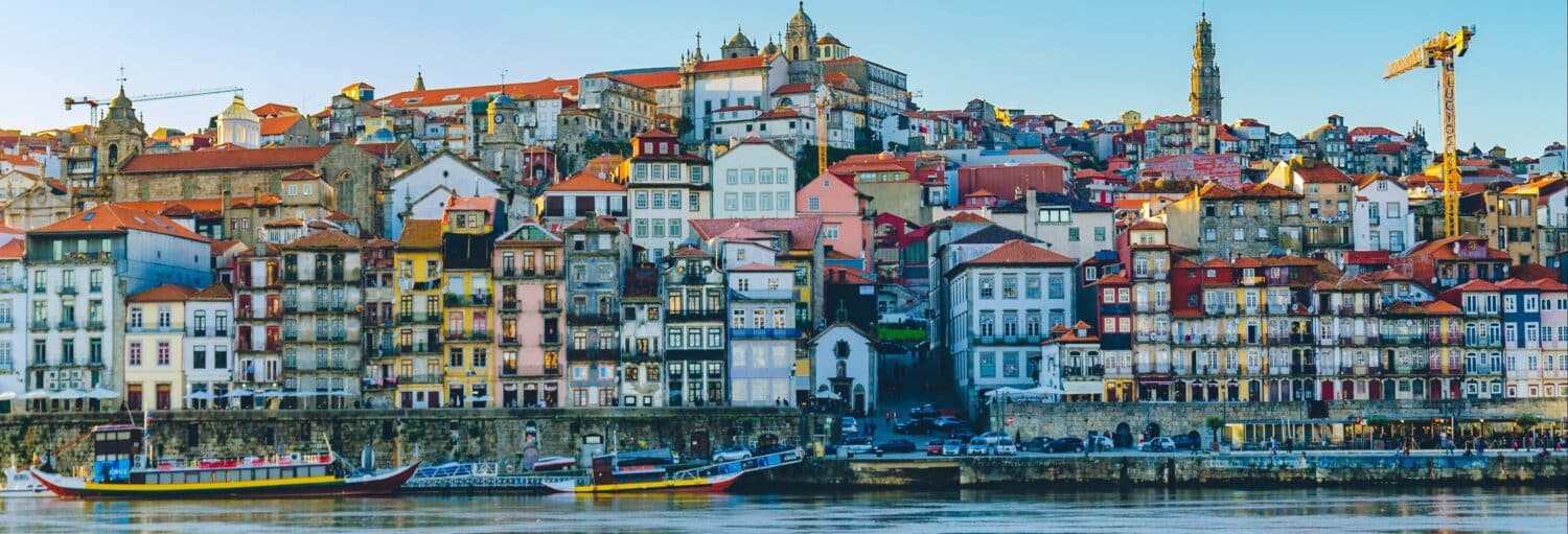 Prédios coloridos se alinham em uma encosta acima de um rio com barcos em Porto, Portugal, sob um céu azul claro. A foto é capa do post Tour pelo Porto medieval e Ribeira.