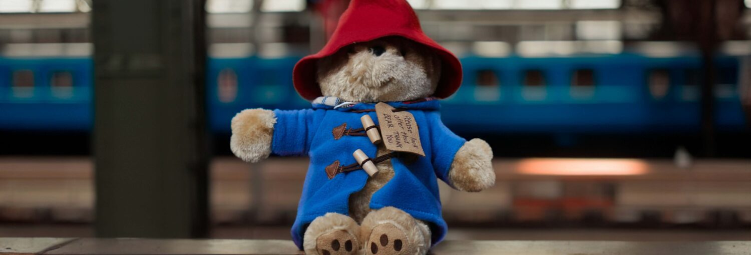 Um ursinho de pelúcia marrom com um casaco azul e chapéu vermelho está sentado em uma saliência, com um trem visível ao fundo.