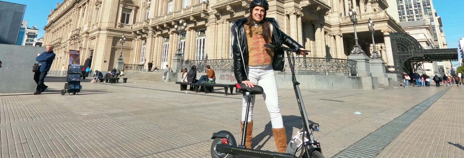 Uma pessoa usando capacete está com uma scooter elétrica em frente a um edifício histórico em uma rua da cidade. Imagem para ilustrar post sobre tour de patinete elétrico por Buenos Aires.