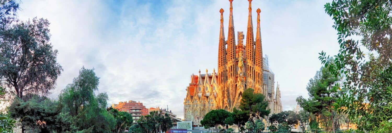 A basílica da Sagrada Família em Barcelona é mostrada com suas altas torres e fachada intrincada cercada por árvores e edifícios. A imagem ilustra o post sobre tour privado pela Sagrada Família