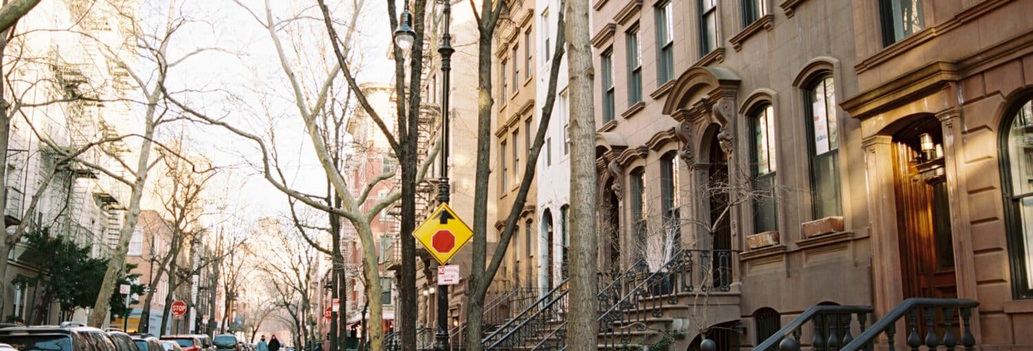 Uma rua da cidade de Nova York com carros estacionados, árvores sem folhas, prédios de arenito e uma placa de trânsito amarela em um dia ensolarado. A foto é capa do post de Tour da série Sex and the City.