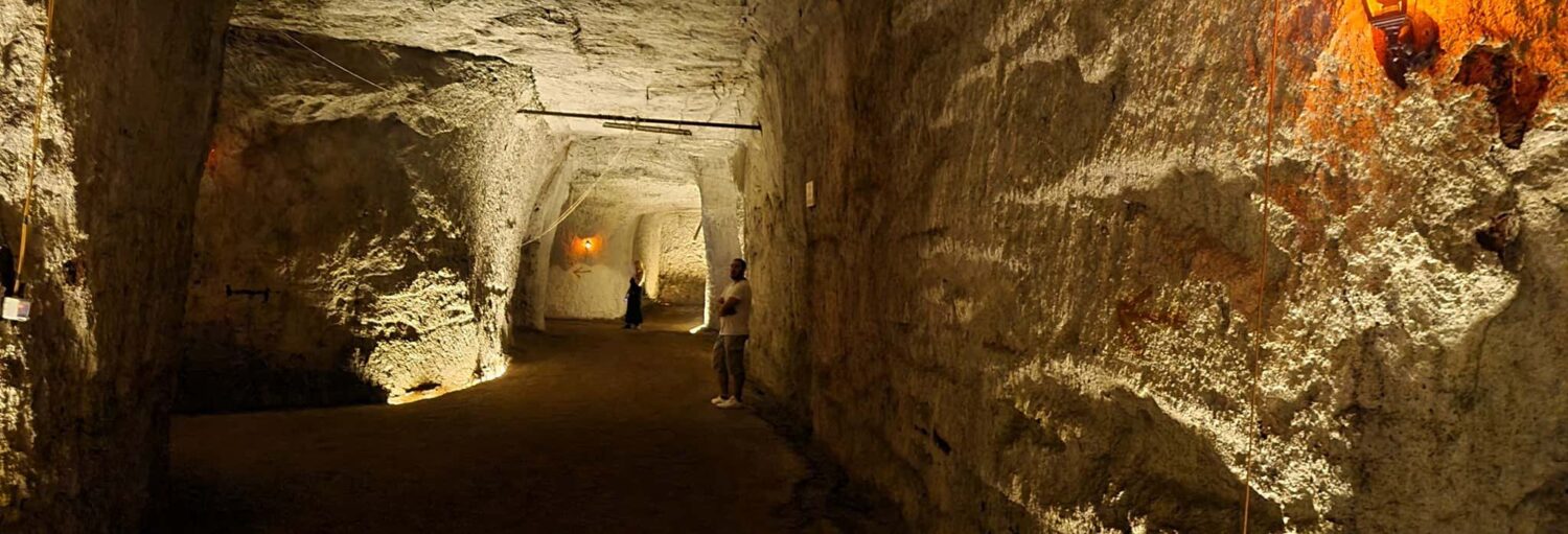 Uma pessoa está dentro de um túnel subterrâneo grande e mal iluminado, com paredes e teto de pedra bruta no Tour pelos túneis da Via Appia e Caffarella