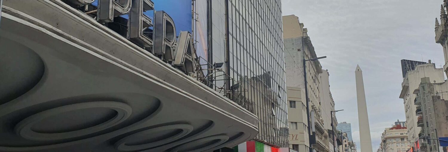 Rua da cidade com prédios altos, uma placa de teatro onde se lê "OPERA" e o Obelisco de Buenos Aires visível ao fundo. Imagem para ilustrar post sobre Tour do tango pela avenida Corrientes.