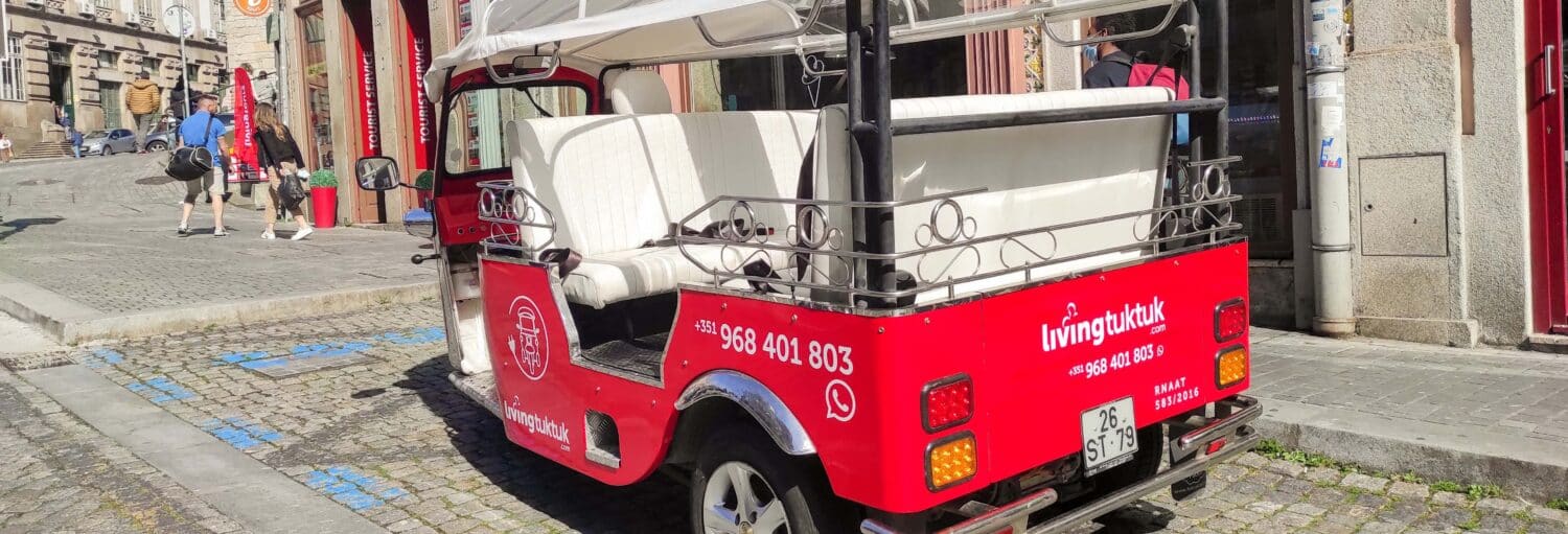 Um tuk-tuk vermelho ao ar livre com assentos brancos está estacionado em uma rua de paralelepípedos perto da entrada de um prédio. A foto é capa do post do Tour de tuk tuk pelo Porto + Cruzeiro das seis pontes.