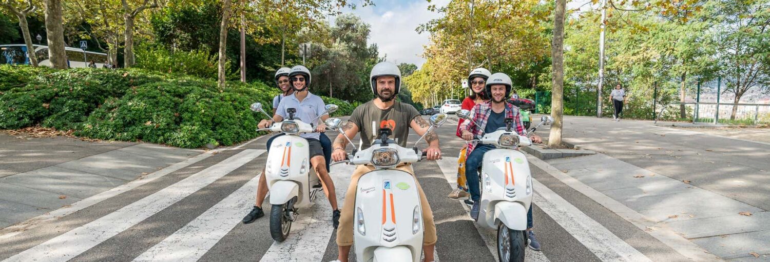 Cinco pessoas usando capacetes estão sentadas em scooters brancas, paradas em uma faixa de pedestres em uma rua arborizada. A imagem ilustra o post sobre tour de vespa por Barcelona