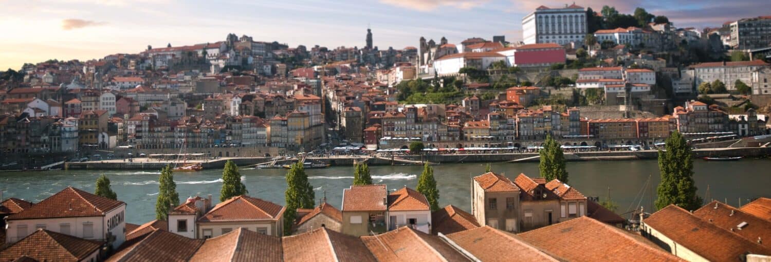 Vista de uma cidade com edifícios de telhados vermelhos ao lado de um rio, com colinas e mais edifícios ao fundo, sob um céu azul. A foto é capa do post do Tour por Vila Nova de Gaia e suas caves.
