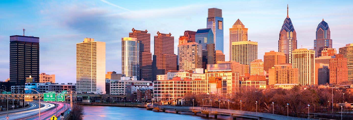 Horizonte da cidade de Filadélfia ao pôr do sol com edifícios e um rio em primeiro plano. A foto é capa do Tour de 2 dias por Washington e Filadélfia.