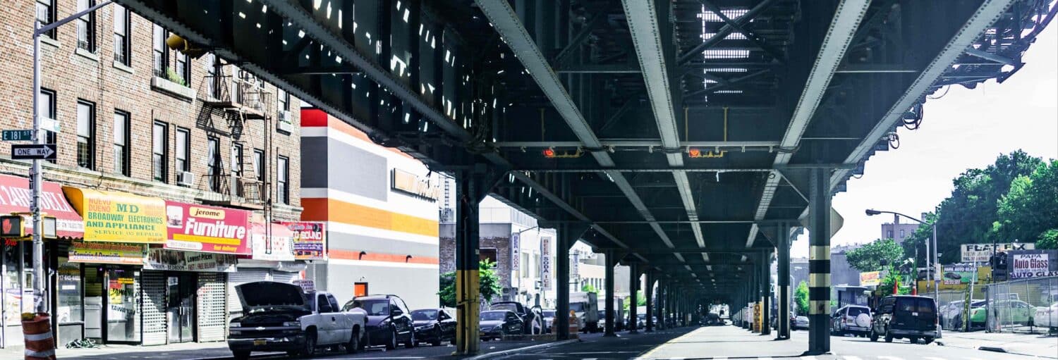 Uma rua da cidade passa sob um trilho elevado de metrô, com carros estacionados e vitrines de lojas em ambos os lados da rua. A foto é capa do post Visita guiada pelo Bronx.