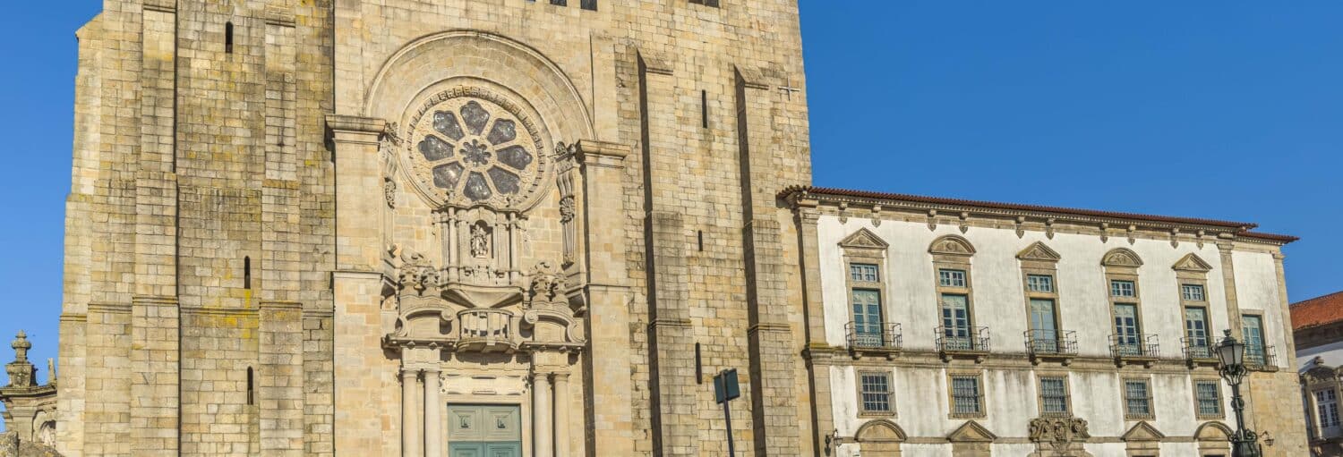 Grande igreja de pedra com uma rosácea detalhada e entrada em arco ao lado de um edifício com várias janelas, sob um céu azul claro. A foto é capa do post da Visita guiada pela catedral do Porto.