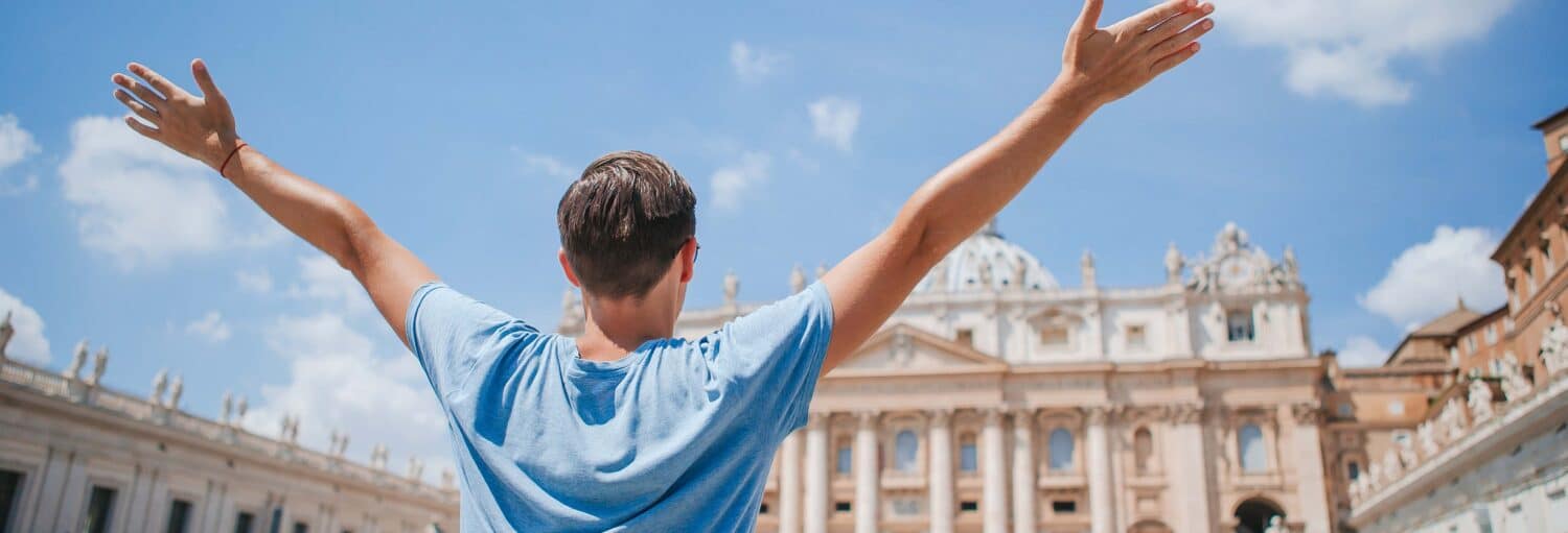 Pessoa com os braços levantados de frente para um grande edifício histórico sob um céu azul com nuvens na Oferta: Vaticano + Coliseu, Fórum e Palatino