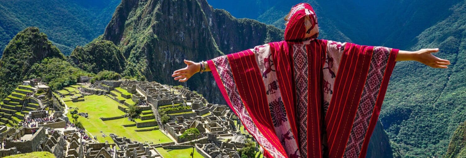 Uma pessoa vestindo roupas tradicionais vermelhas, com os braços estendidos, observa as ruínas de Machu Picchu, no Peru na Excursão de 2 dias a Machu Picchu com ingressos