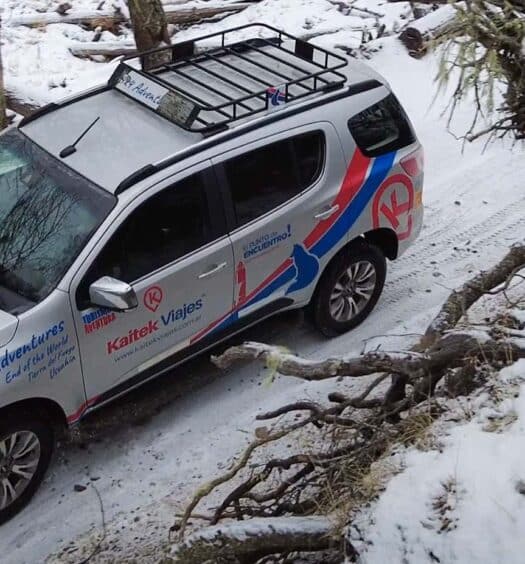 Um SUV prateado com gráficos vermelhos e azuis dirige em uma estrada de terra coberta de neve por uma floresta nevada com árvores caídas. Imagem para ilustrar post sobre o tour de 4x4 pelos lagos Escondido e Fagnano + Estância La Carmen.
