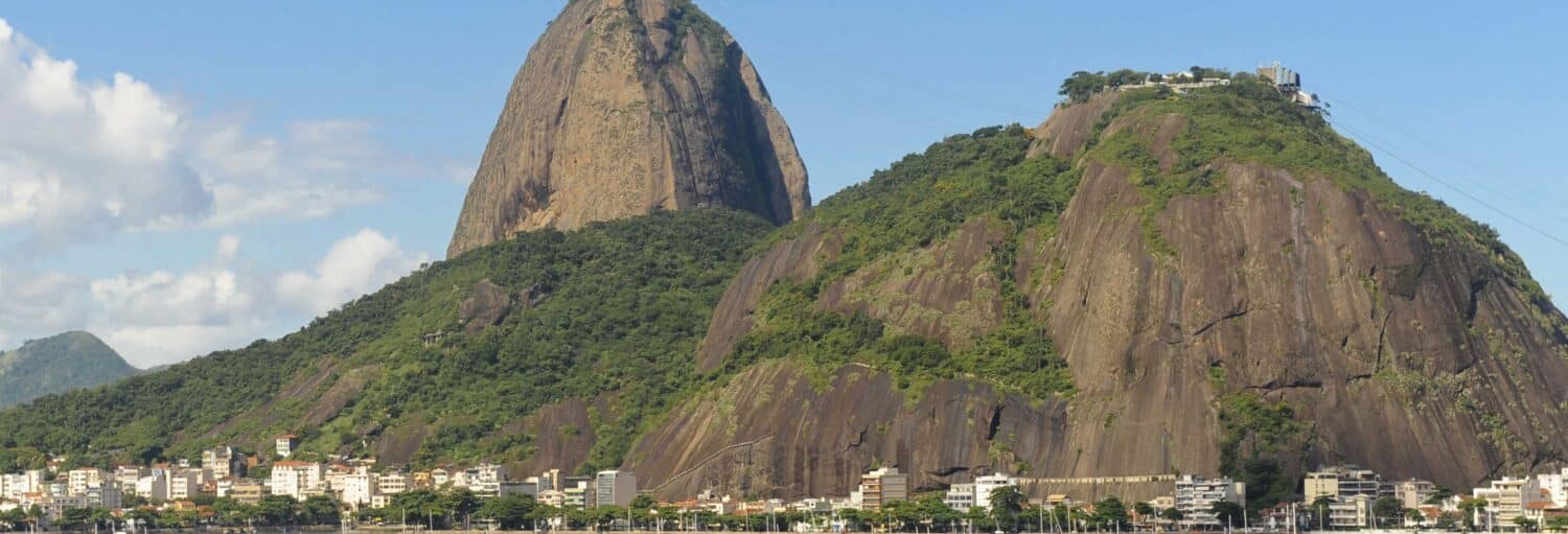 Grande montanha rochosa com vegetação verde exuberante e edifícios em sua base sob um céu azul com nuvens dispersas. A imagem ilustra o post sobre Aluguel de barco com capitão no Rio.