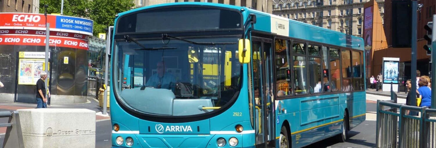 Um ônibus urbano azul da Arriva está parado em um cruzamento urbano em um dia ensolarado com pessoas por perto. A foto é capa do post do ônibus entre o Aeroporto John Lennon e Liverpool.