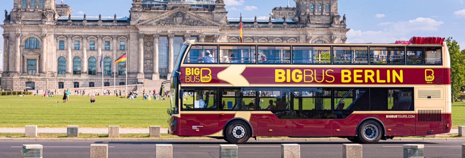 Um ônibus de turismo vermelho e amarelo de dois andares com o nome "Big Bus Berlin" passa em frente a um edifício histórico com uma cúpula de vidro. Imagem para ilustrar post sobre ônibus turístico de Berlim, Big Bus + passeio de barco.