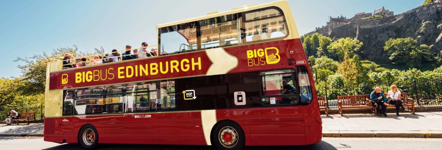Um ônibus de turismo Big Bus Edinburgh de dois andares com a parte superior aberta passa por um muro de pedra; as pessoas sentam-se em bancos próximos. A foto é capa do post do Ônibus turístico de Edimburgo.