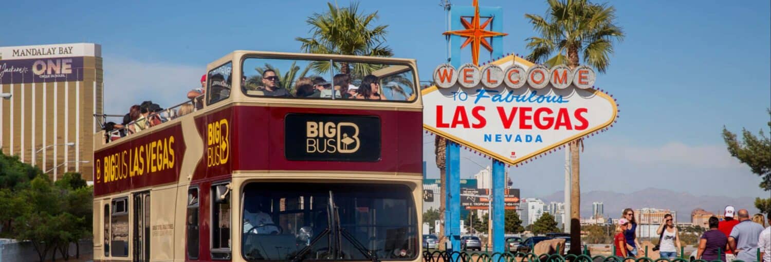 Um ônibus de turismo de dois andares passa pela placa Welcome to Fabulous Las Vegas com palmeiras e hotéis ao fundo. Imagem para ilustrar post sobre ônibus turístico de Las Vegas.