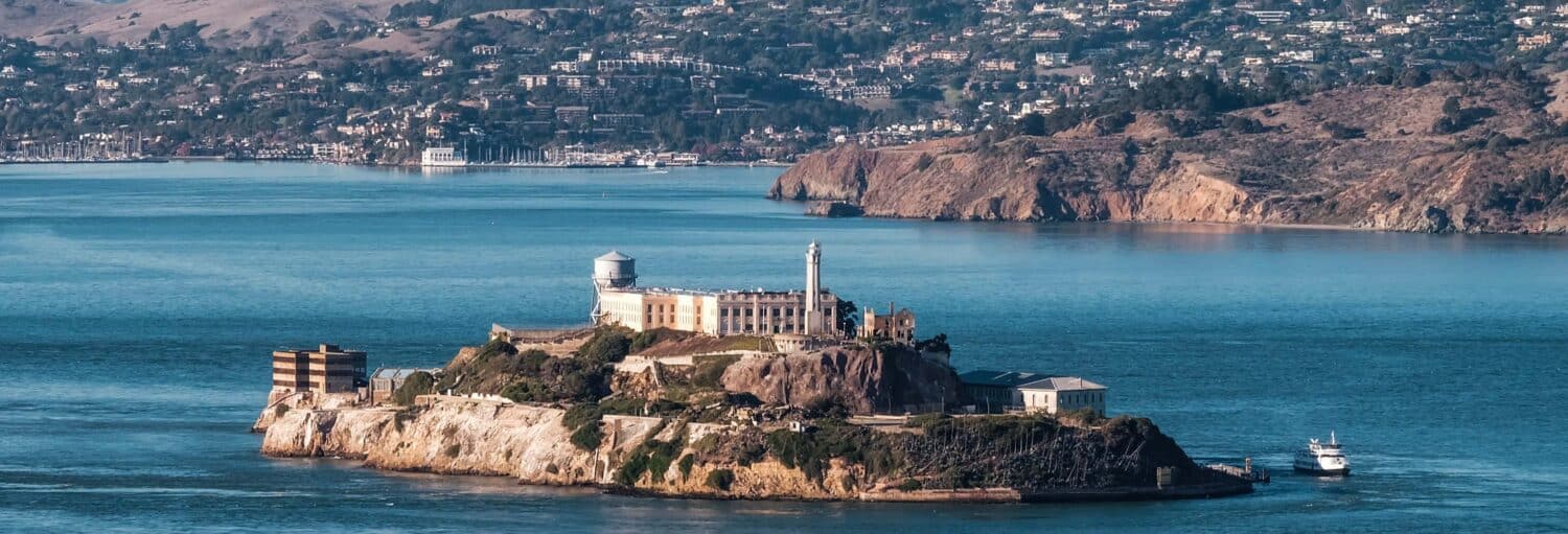 Uma ilha com edifícios históricos e um farol, cercada por água, com colinas e casas ao fundo. - Ônibus turístico de São Francisco + Ingresso de Alcatraz