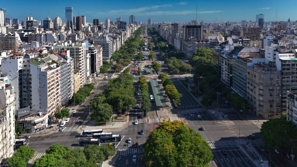 Uma ampla avenida urbana repleta de árvores, prédios altos em ambos os lados e veículos na estrada sob um céu claro, na Avenida 9 de Julio em Buenos Aires.