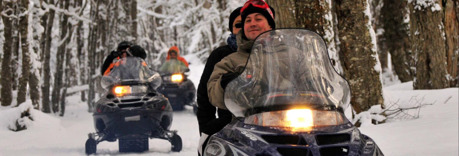 Três pessoas andam de snowmobiles em uma fila por uma floresta nevada com árvores nuas em um dia de inverno. Imagem para ilustrar post sobre a aventura noturna de moto de neve, trenó e raquetes.
