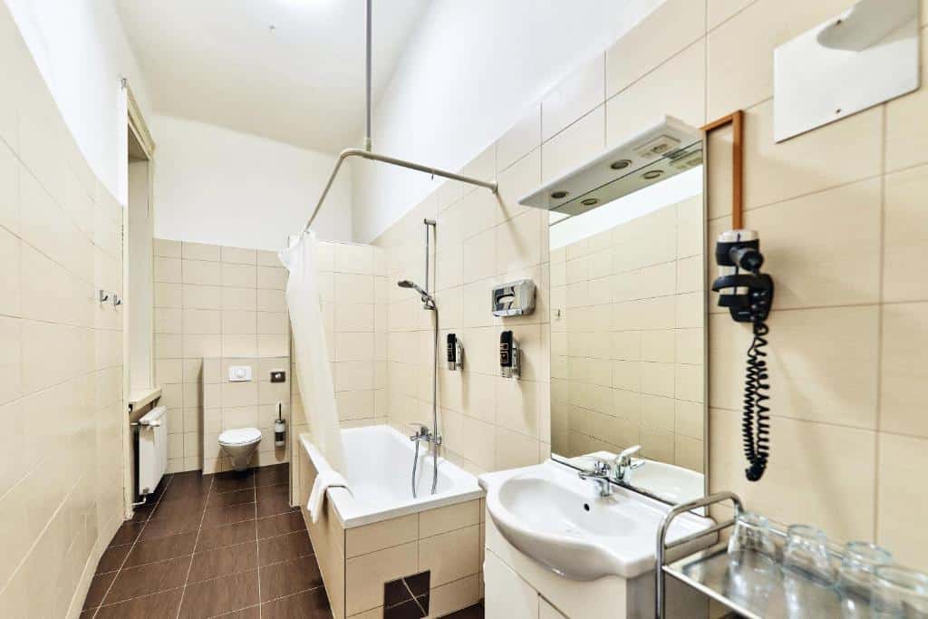 Um banheiro azulejado com banheira, chuveiro, vaso sanitário, pia, secador de cabelo de parede e vários copos de vidro em uma bandeja de metal, no Hotel Pension Baron am Schottentor.