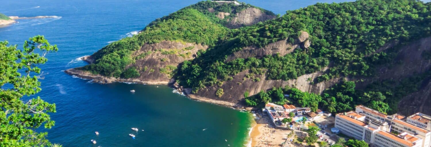 Vista aérea de uma área costeira com colinas verdes, água azul, uma praia de areia e edifícios próximos à costa. A imagem ilustra o post sobre Barco pelas Ilhas Cagarras + Praia Vermelha.