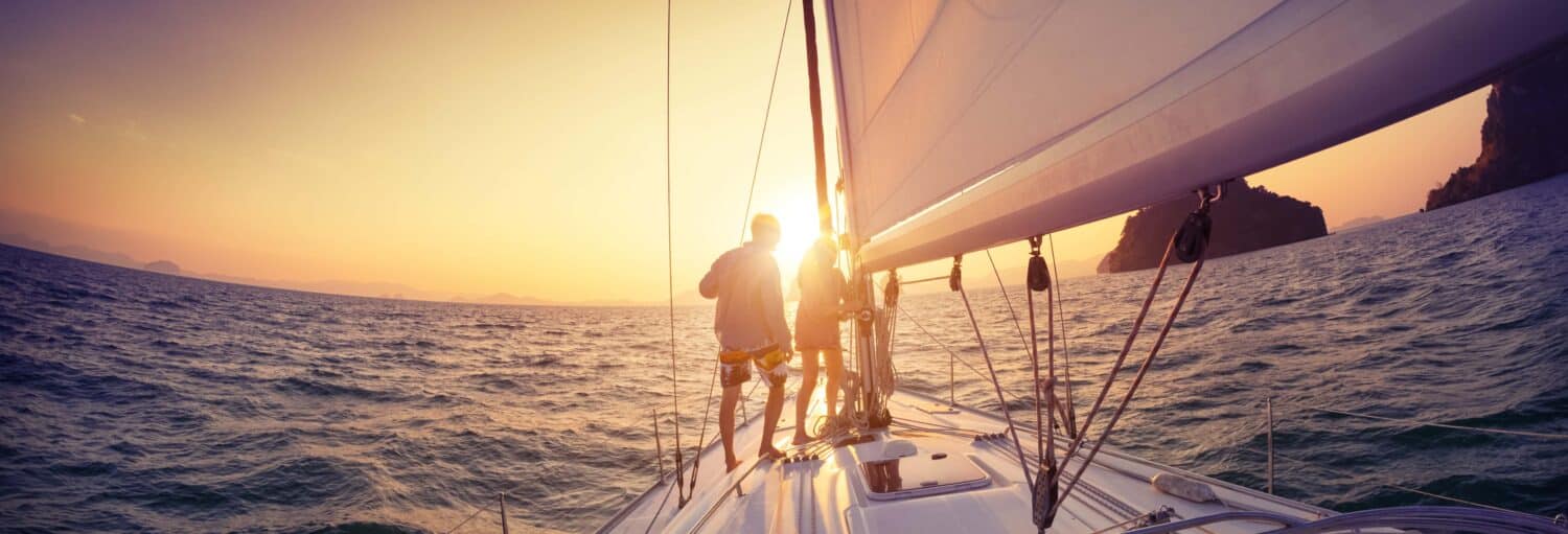 Duas pessoas desfrutam de um passeio de barco ao entardecer pelo Rio, em pé em um veleiro enquanto o sol se põe em uma terra distante.