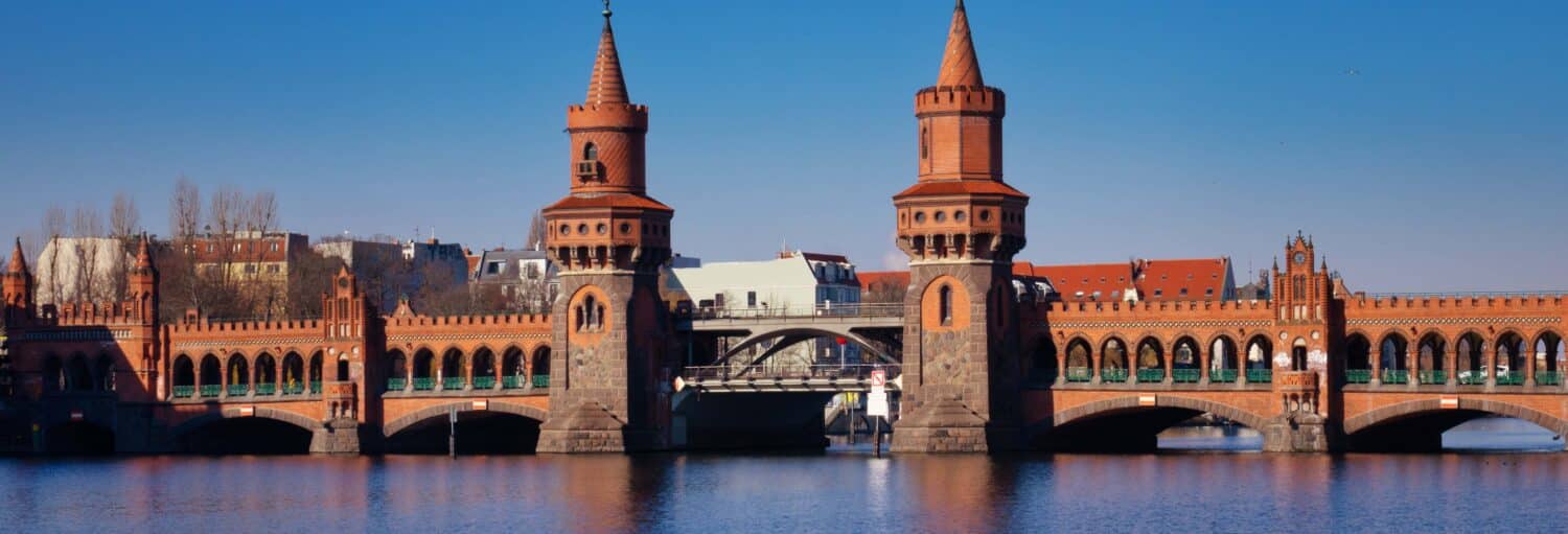 Uma ponte de tijolos vermelhos com duas torres atravessa um rio sob um céu azul claro, com edifícios ao fundo. Imagem para ilustrar post sobre barco turístico de Berlim.