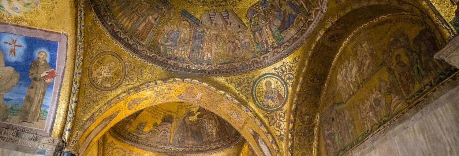 Arcos de mosaico dourado e afrescos religiosos pintados decoram o teto e as paredes do interior de uma igreja histórica. A imagem ilustra o post sobre Tour pela Basílica de São Marcos e Pala D’Oro.
