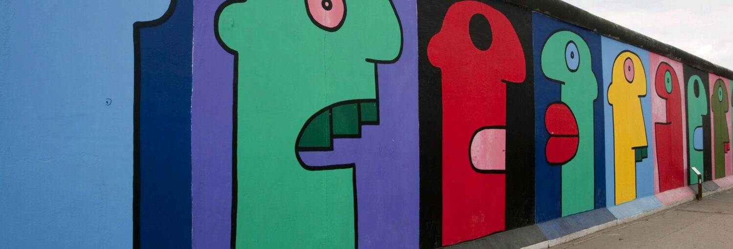 Rostos abstratos coloridos pintados em uma fileira em um segmento do Muro de Berlim, vistos de um ângulo. Imagem para ilustrar post sobre visita guiada pela Berlim alternativa.