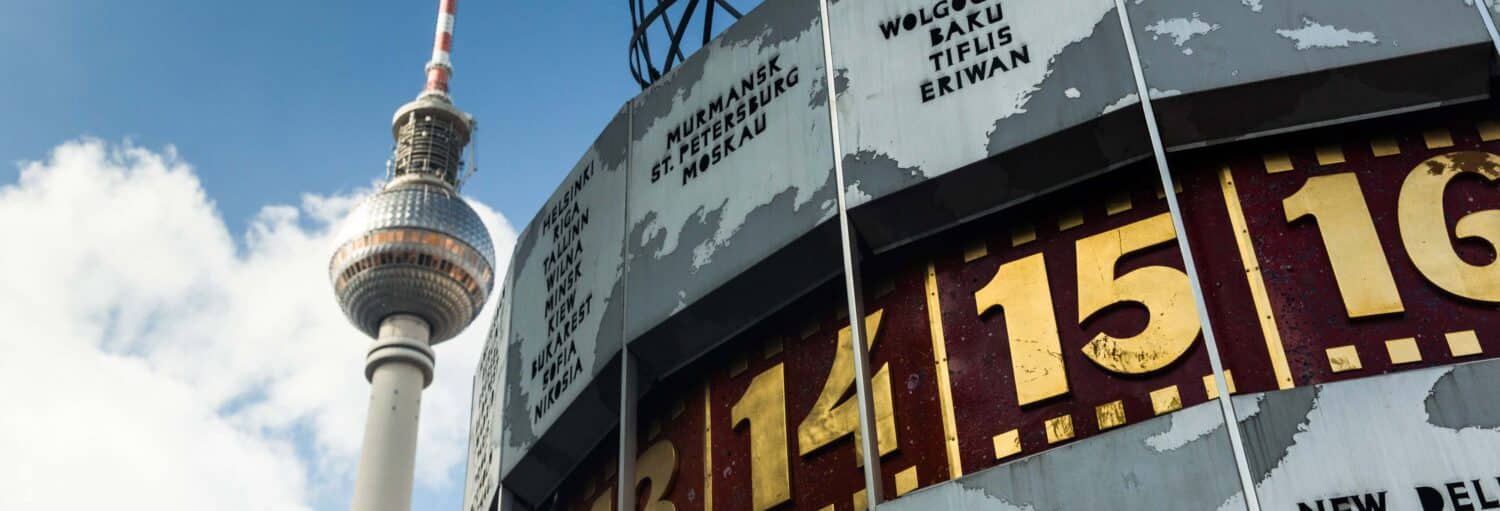 Close-up do Relógio Mundial de Berlim mostrando números e nomes de cidades, com a Torre de TV de Berlim ao fundo contra o céu. Imagem para ilustrar post sobre tour do Muro de Berlim.