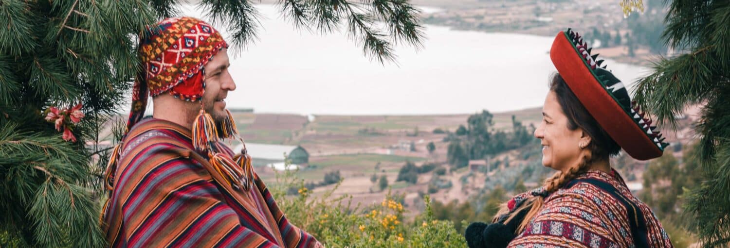 Duas pessoas com roupas andinas tradicionais estão ao ar livre, de frente uma para a outra e sorrindo, com uma paisagem ao fundo no Casamento inca. Case seguindo o ritual tradicional!