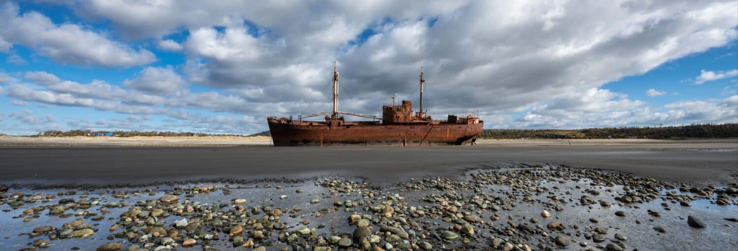 Um navio enferrujado e abandonado fica em uma praia de areia sob um céu nublado com rochas espalhadas em primeiro plano. Imagem para ilustrar post sobre excursão ao Cabo San Pablo e Estância Pirinaica.