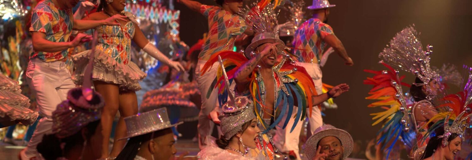 Um grupo com trajes coloridos e chapéus prateados dança no palco do animado Espetáculo Roxy Dinner Show.