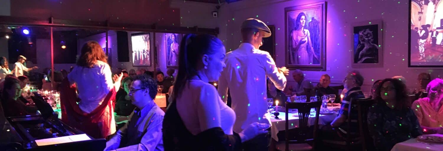 Os artistas dançam e tocam piano em um restaurante mal iluminado, com clientes sentados em mesas e obras de arte nas paredes. A imagem ilustra o post sobre Jantar com ópera e zarzuela no La Castafiore.