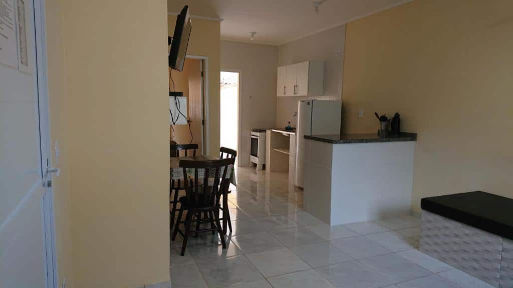 Uma pequena cozinha e área de jantar com mesa, cadeiras, geladeira, paredes de cor neutra e piso de azulejo, nos Chalés Mallumar na Maranduba.