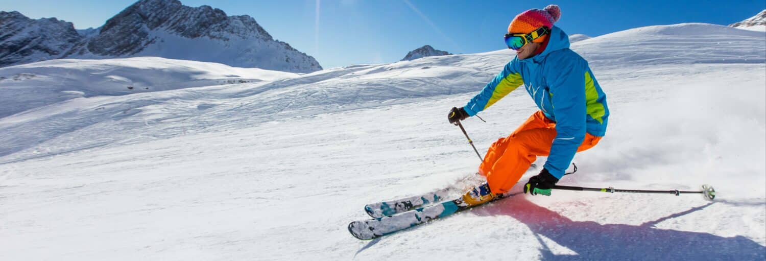 Um esquiador usando um equipamento brilhante faz uma curva em uma pista de neve com montanhas e céu azul ao fundo. Imagem para ilustrar post sobre excursão a Grindelwald com aula de esqui.