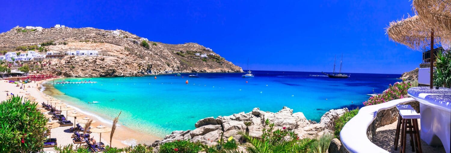 Uma praia ensolarada com água azul-turquesa transparente, colinas rochosas, espreguiçadeiras e barcos ancorados na baía sob um céu azul brilhante. A imagem ilustra o post sobre Barco pelo sul de Mykonos com almoço.
