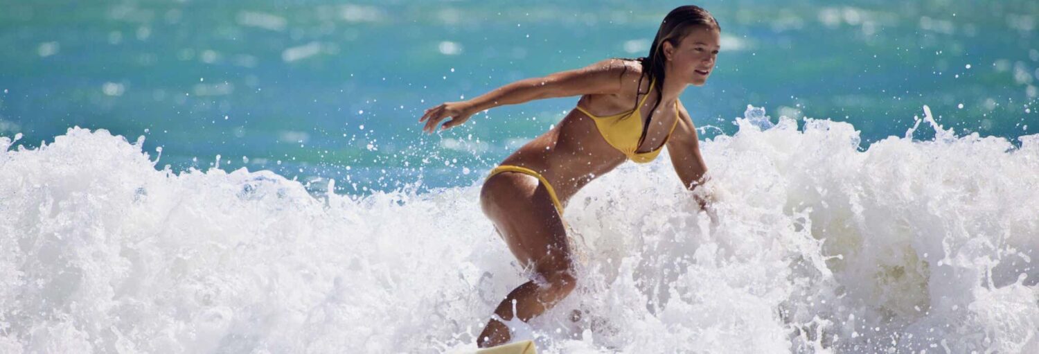 Uma mulher de biquíni amarelo surfa em ondas brancas, fazendo um curso de surfe em Miami sob a luz do dia.